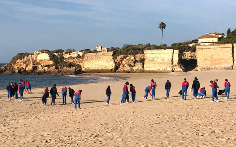 Escolares participan en una pasada limpieza de la playa con el monumento al fondo / FOTO: Ayto.